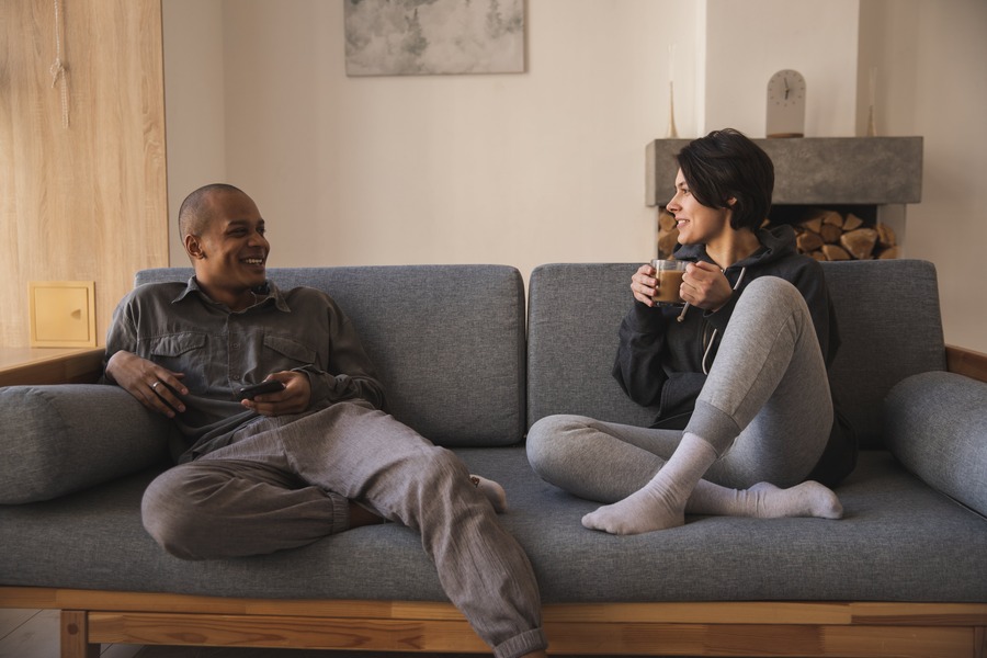 Couple sitting on the couch talking and smiling, the woman holding a cup of coffee and the man holding his smartphone.