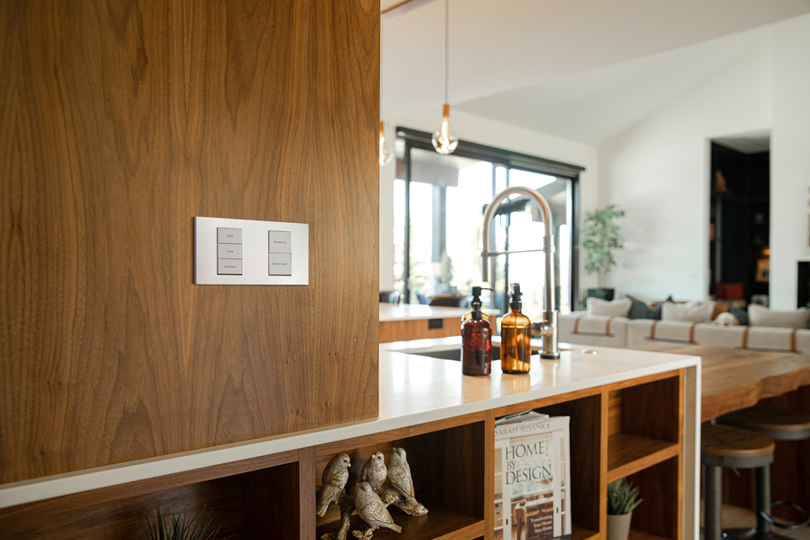 A Control4 wall keypad in the foreground with a kitchen island and sink in the background.