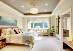 A luxury bedroom with recessed lighting on the ceiling and a large window with open curtains on the back.