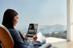 A person sitting in a chair looking out over a cityscape, holding an iPad displaying the Savant home automation platform.